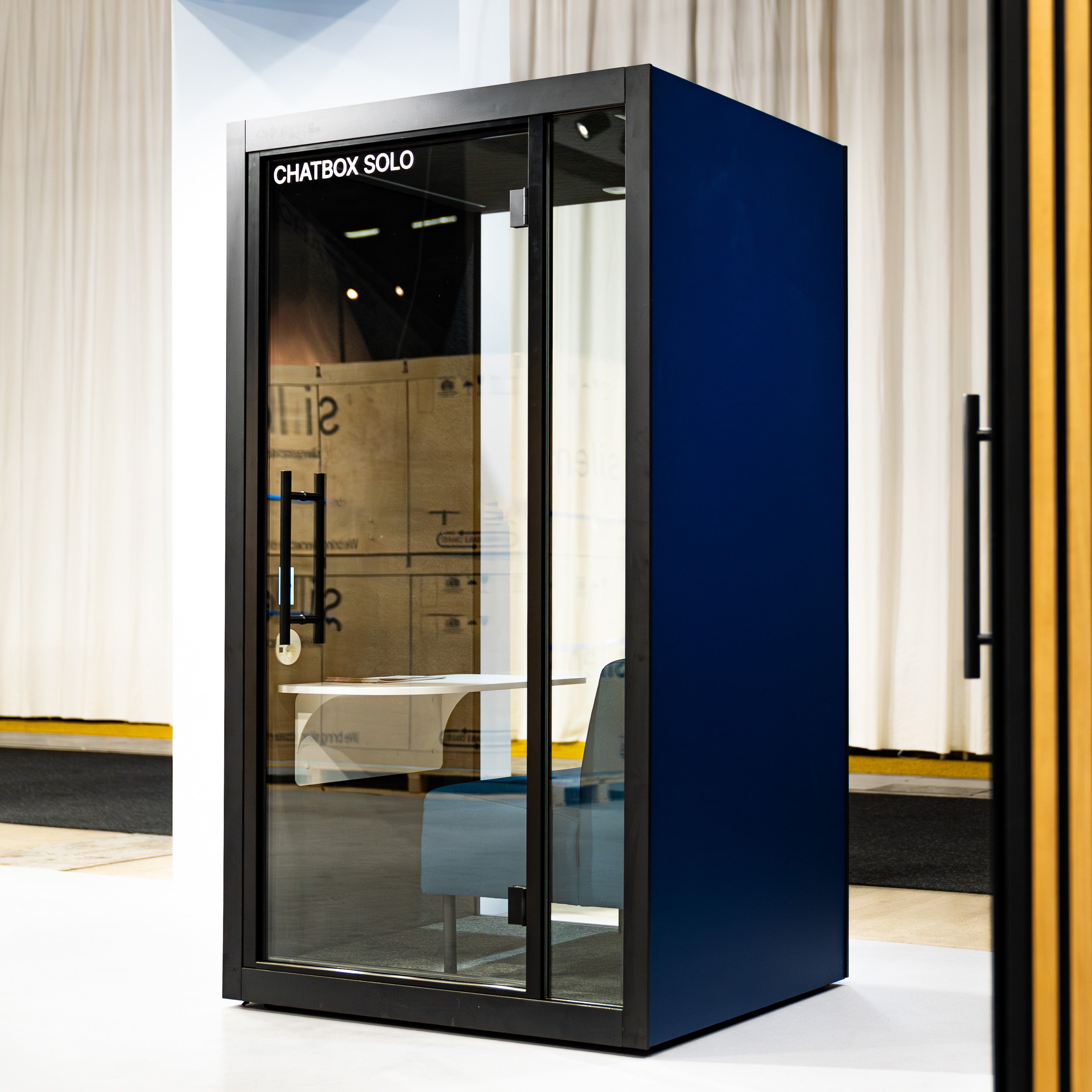 Chatbox Solo Phone Box mit blauer Rueckwand und grosser Glasfront – moderne Akustikkabine mit integriertem Tisch und Sitzmoebel fuer ruhiges Arbeiten im offenen Buero