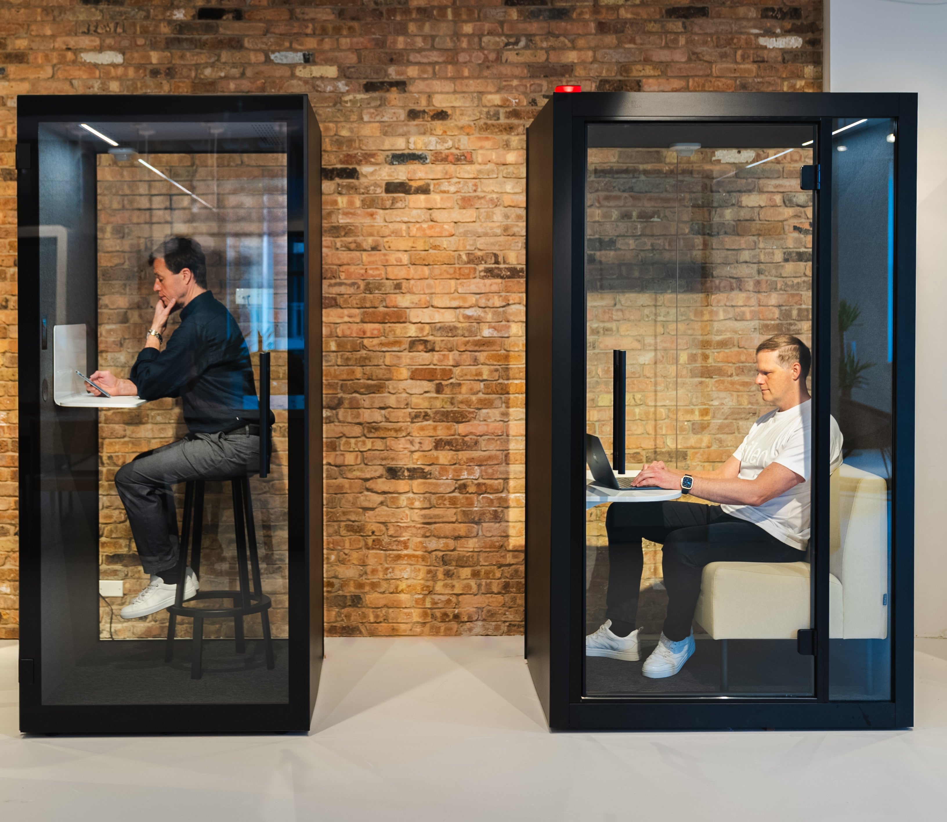 Zwei Chatbox Solo Phone Boxen in modernem Buero mit Ziegelwand – links stehender Arbeitsplatz mit Barhocker, rechts Sitzgelegenheit mit Tisch fuer konzentriertes Arbeiten in akustisch geschuetztem Raum